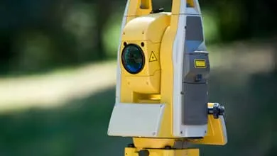 Close-up of a yellow and white surveying total station, outdoors, with a blurred green background.