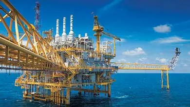A large yellow and white offshore oil rig stands in the ocean under a clear, sunny sky.