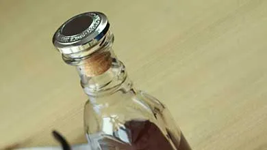 Close-up of a glass bottle with a cork sealed by a round silver decorative cap, set on a light wood-grain surface.