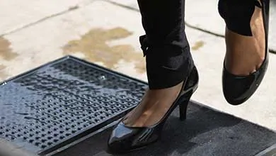 A person in black pants and shiny black heels steps onto a mat beside a wet metal grate on the sidewalk.