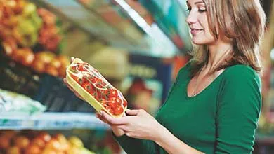 A woman in a green shirt holds a package of cherry tomatoes in a grocery store produce section.