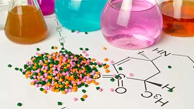 Green, orange, and pink plastic pellets on a white surface with chemical structure drawings; behind them are glass flasks filled with colorful liquids.