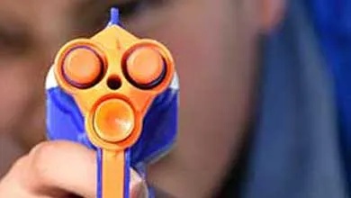 Close-up of a blue and orange toy gun pointed at the camera, with a boy aiming in the blurred background.