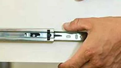 Two hands adjust a metal drawer slide on a white drawer, with the silver track and mounting hardware visible against white cabinetry.