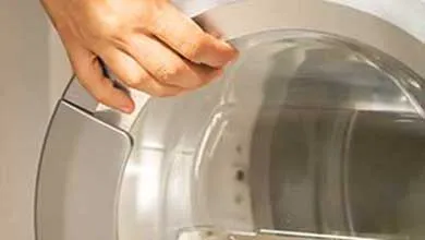 A hand holds the circular door of a silver front-loading washing machine or dryer, opening or closing the transparent glass door.