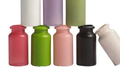 Nine small cylindrical bottles in different colors are arranged in two rows on a white background. The top row has three upright bottles: white, lavender, and olive green. The bottom row has five upright bottles—coral red, lime green, peach, black—and one off-white speckled bottle lying on its side.
